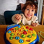child, toddler, smiling, playing, toy, fishing_game, table, indoor, adult, fridge, white_shirt, wooden_table, happy, face, hand, colorful, game, furniture, person, hair