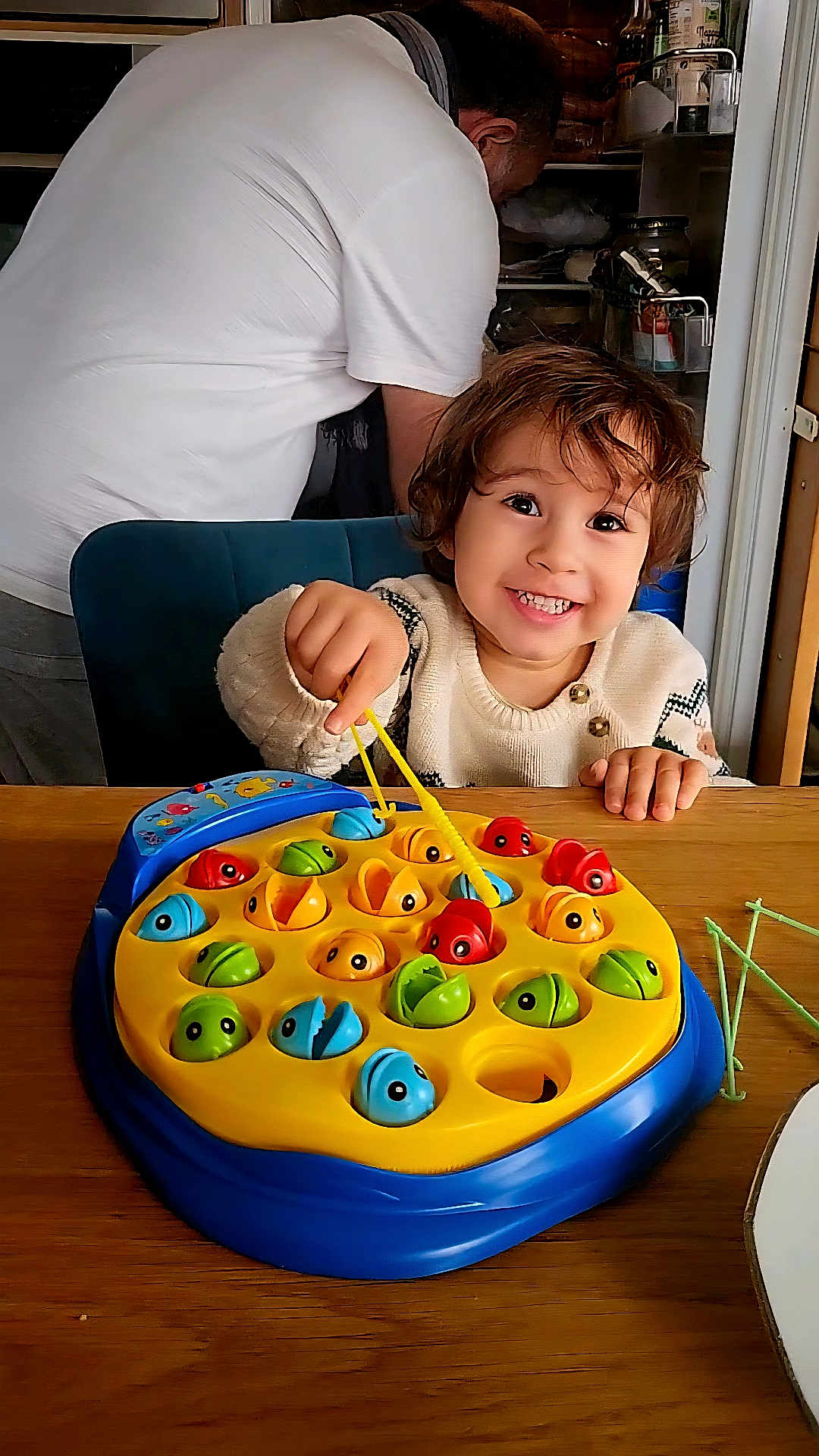 Aidan a rejoint le concours — aidez-le/la à gagner de superbes lots ! child, toddler, smiling, playing, toy, fishing_game, table, indoor, adult, fridge, white_shirt, wooden_table, happy, face, hand, colorful, game, furniture, person, hair