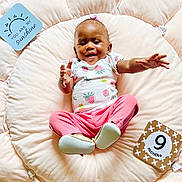 Shevon is registered to the contest to win money with this photo: baby, child, clothing, cute, face, feet, hands, happy, indoors, infant, milestone, milestone_card, person, pink_pants, plush, portrait, smiling, soft_cushion, strawberry_shirt, wood_floor
