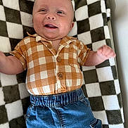 Liam is registered to the contest to win money with this photo: baby, smiling, checkered_shirt, denim_pants, blanket, plaid, happy, child, cute, infant, clothing, portrait, lying_down, indoor, face, hands, buttoned_shirt, blue_pants, texture, cozy