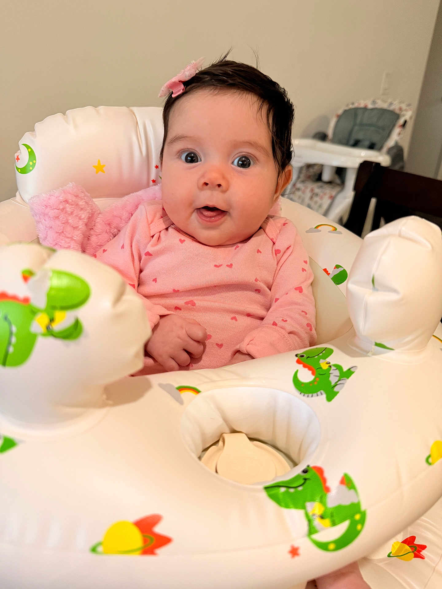 Luna is registered to the contest to win money with this photo: baby, infant, pink_clothing, onesie, inflatable_ring, dinosaur_print, bow, hair_accessory, wide_eyes, surprised_expression, indoor, high_chair, table, child, cute, seated, soft_toy, playful, colorful, portrait