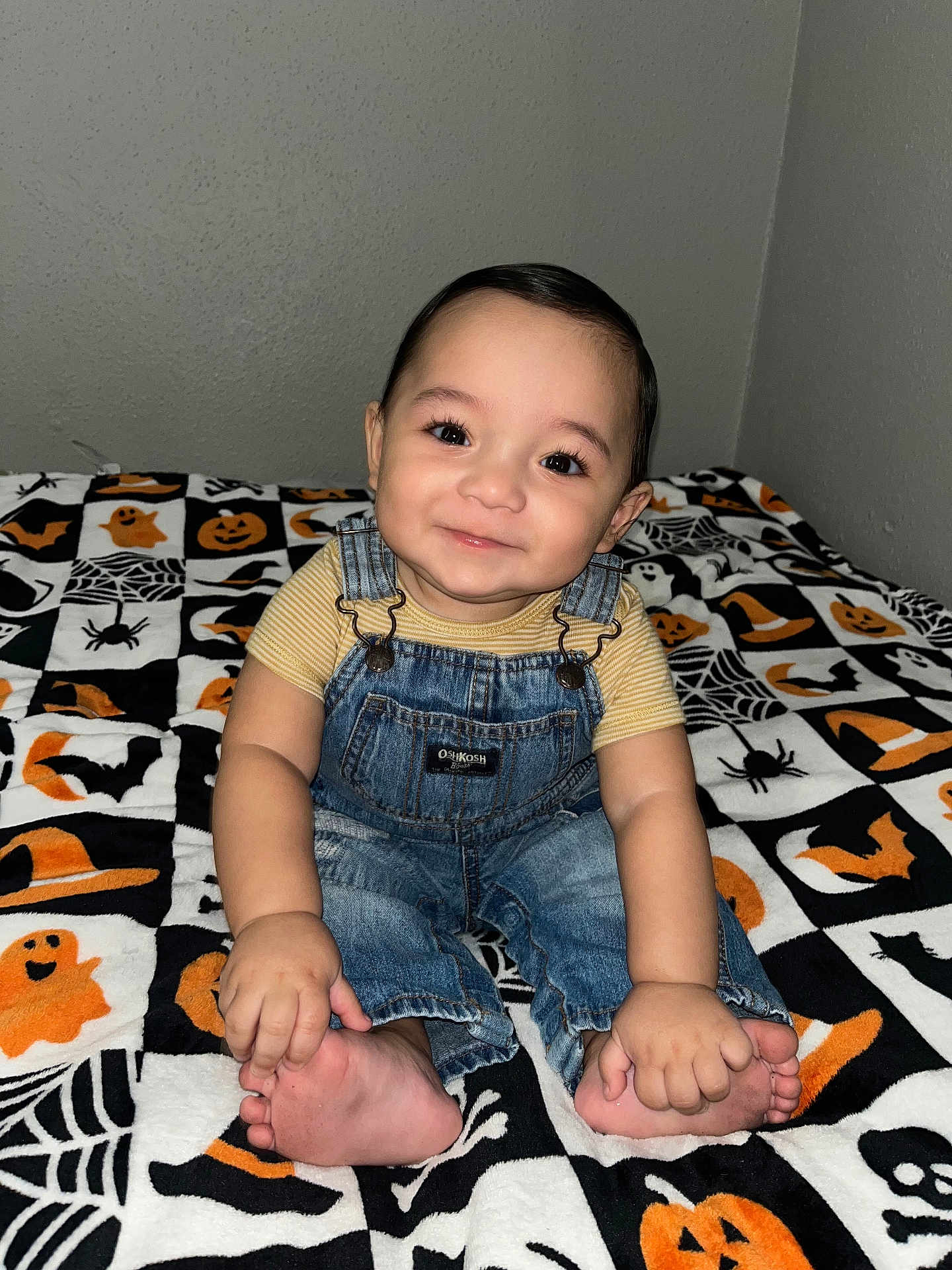Jack is registered to the contest to win money with this photo: baby, child, smile, denim_overalls, yellow_shirt, barefoot, hands, feet, bed, blanket, halloween_theme, pumpkins, spiders, ghosts, black, orange, white, indoor, cute, portrait