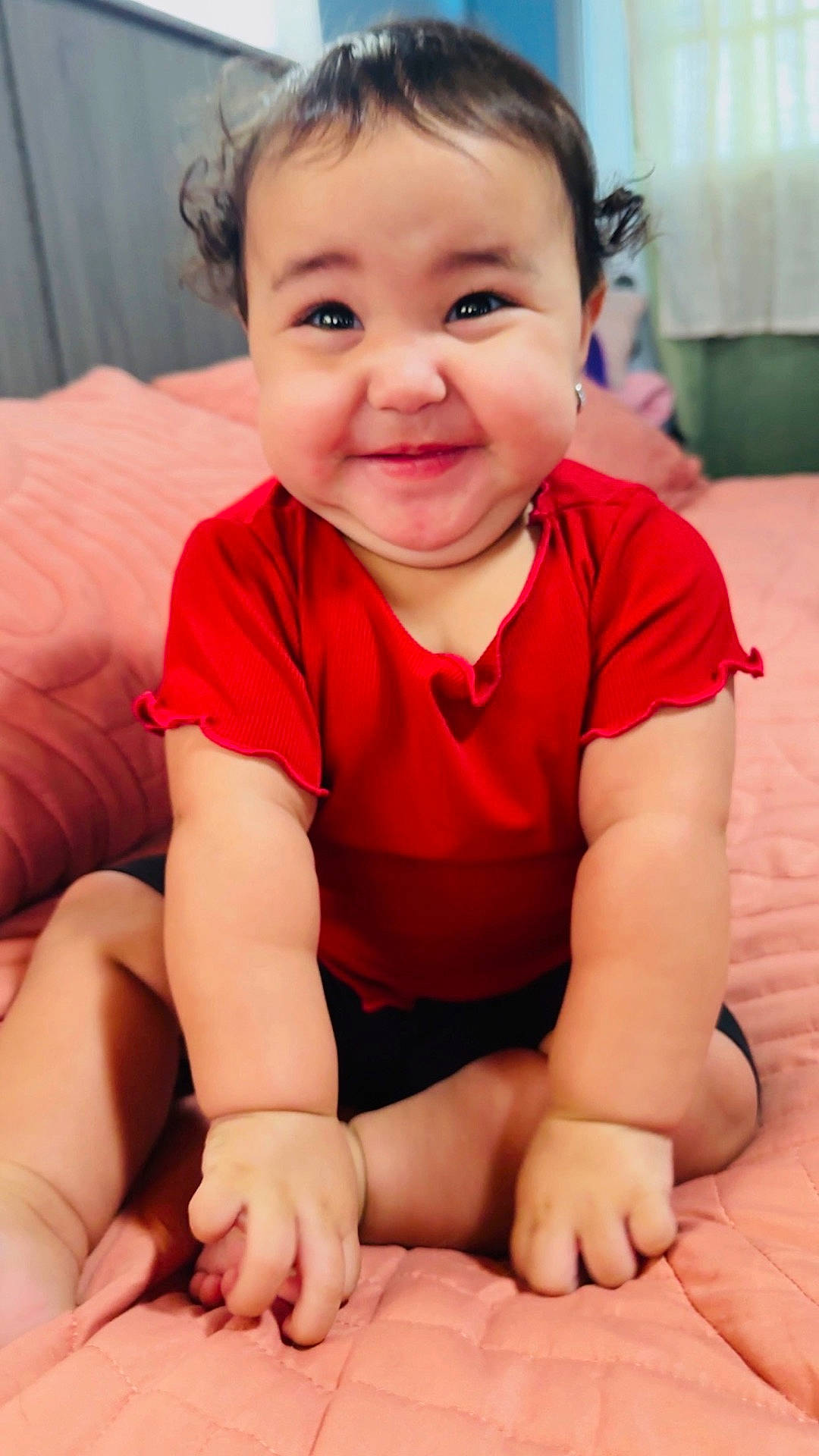 Josiris Isarel is registered to the contest to win money with this photo: baby, bed, cheeks, child, curly_hair, cute, eyes, happy, indoors, infant, mouth, nose, person, portrait, quilt, red_shirt, sitting, skin, small_hands, smiling