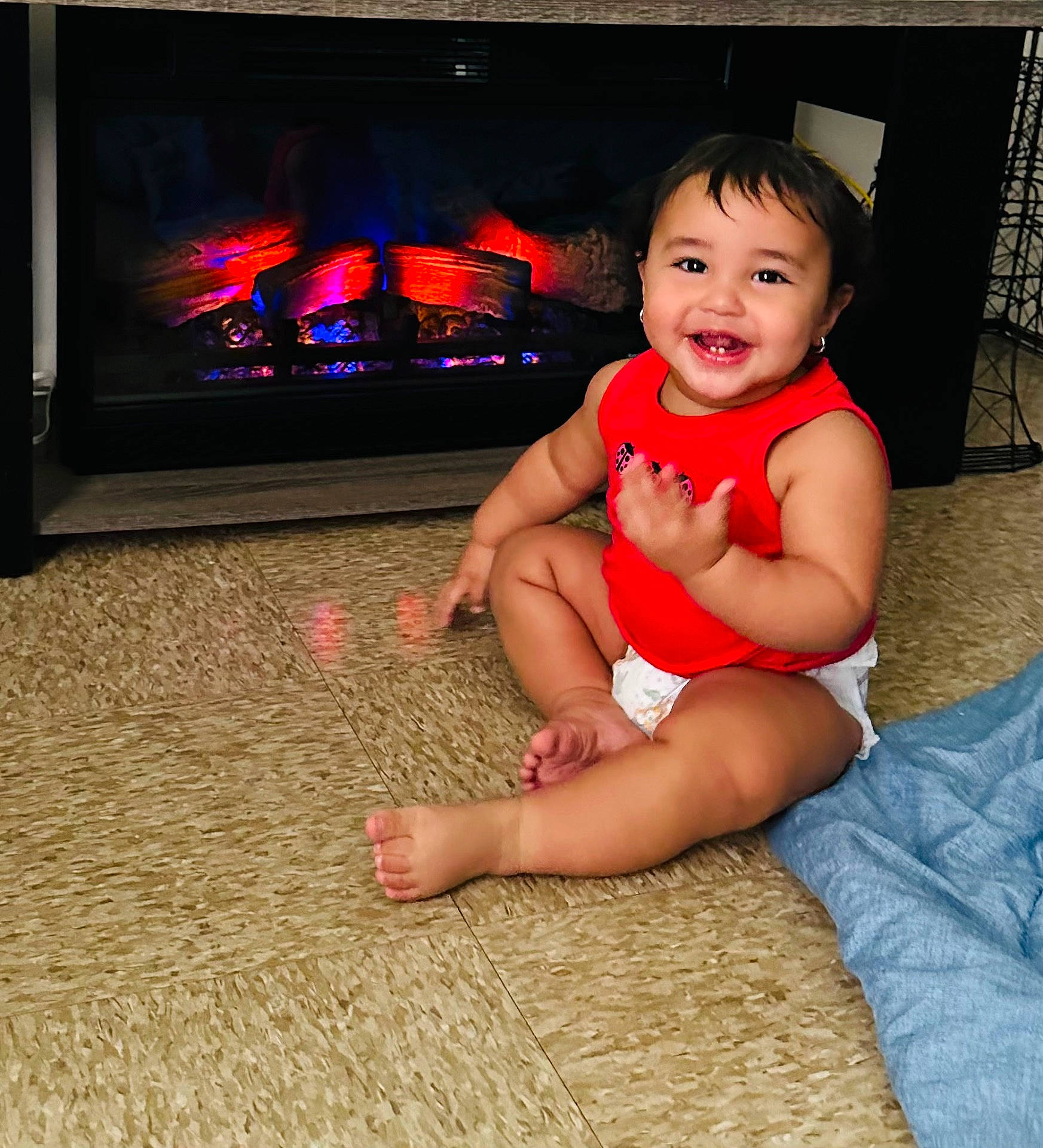 baby, blanket, child, cozy, cute, diaper, electric_fireplace, face, floor, happy, home, indoor, person, red_shirt, sitting, smiling, toddler, toes, warm, young_child
