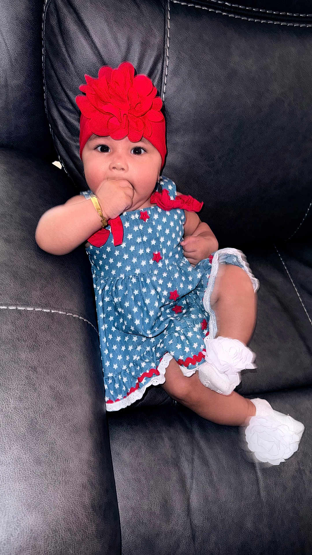 Josiris Isarel is registered to the contest to win money with this photo: baby, infant, child, dress, flower_headband, red_hat, bracelet, sock, ruffle, couch, leather_sofa, sitting, portrait, cute, hand_in_mouth, stars_pattern, white_flowers, indoor, blue_dress, adorable