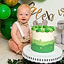 baby, child, cake, birthday, balloons, green, gold, bow_tie, suspenders, plush_toy, elephant, celebration, floor, smiling, happy, party, decorations, white_stand, plant_leaves, indoor