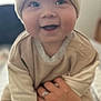 baby, infant, smile, face, hat, knit_hat, beige_clothing, hand, ring, person, cute, happy, indoors, soft_light, portrait, child, warm, cozy, sitting, closeup