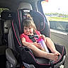toddler, child, car_seat, vehicle_interior, sippy_cup, window, gas_station, greenery, pink_clothing, black_car_seat, seatbelt, cheeky_smile, hair_tie, footwear_free, car_door, daylight, seat, baby, portrait, sitting