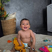 Lisandro a rejoint le concours — aidez-le/la à gagner de superbes lots ! baby, smiling, toy, diaper, yellow_clothing, rug, pillow, basket, plant, indoor, happy, child, playful, floor, cute, sitting, colorful, accessory, home, infant