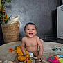 baby, smiling, toy, diaper, yellow_clothing, rug, pillow, basket, plant, indoor, happy, child, playful, floor, cute, sitting, colorful, accessory, home, infant