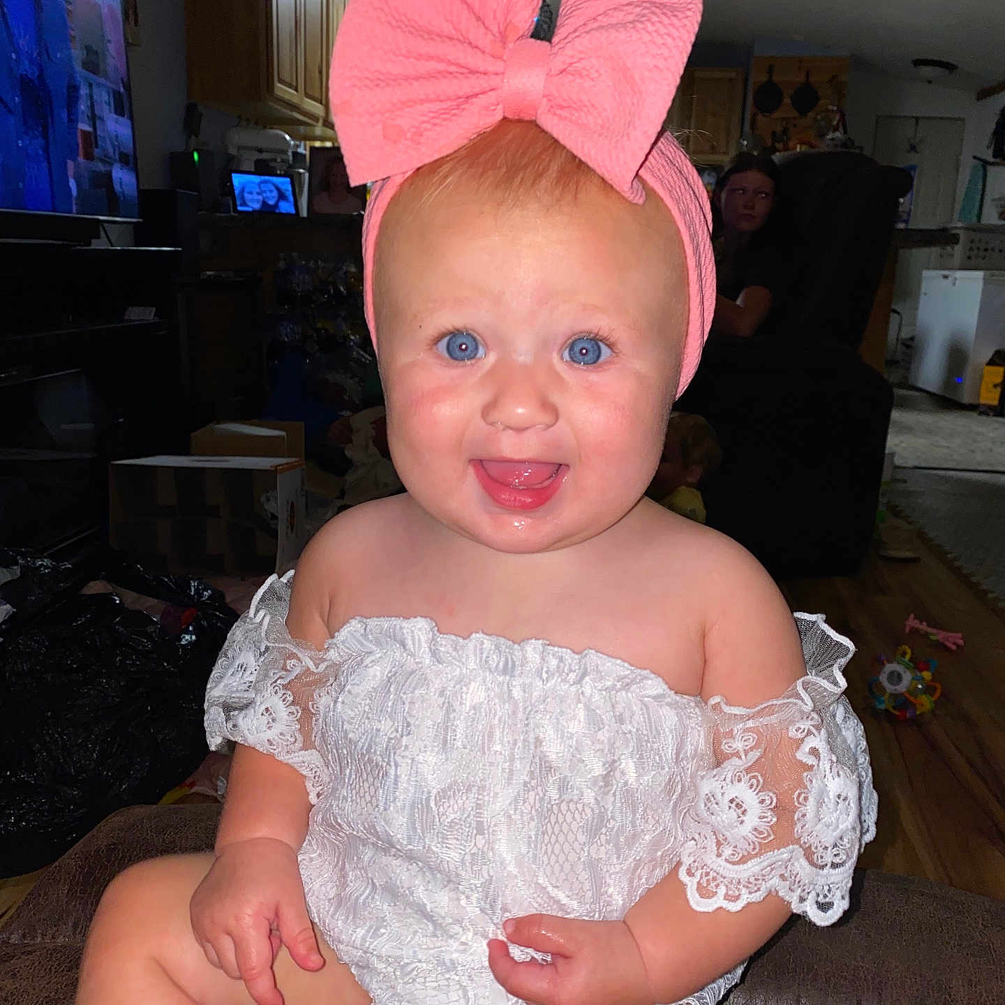 Wrenleigh is registered to the contest to win money with this photo: baby, child, blue_eyes, pink_bow, white_lace_outfit, sitting, smiling, indoor, living_room, couch, furniture, person, television, cabinet, floor, wooden_floor, toy, headband, happy, portrait