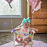 baby, child, infant, smiling, chair, balloon, pink, headband, ribbon, colorful_clothing, wooden_furniture, carpet, window, sunlight, indoor, celebration, happy, cute, portrait, room
