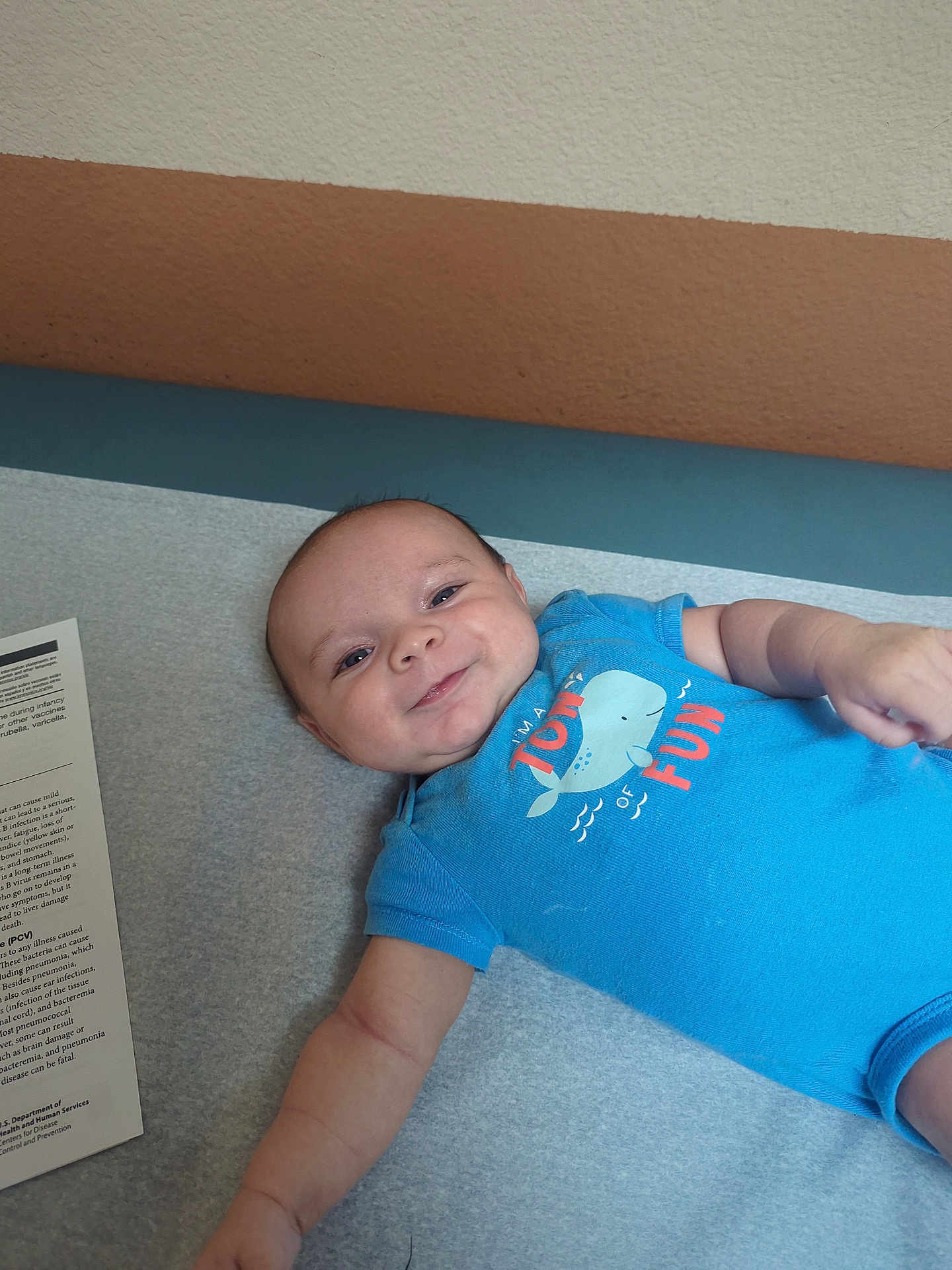 Dominic Sage Colon is registered to the contest to win money with this photo: arm, baby, blue_onesie, child, cute, face, happy, healthcare, indoor, infant, laying_down, medical_table, newborn, paper, person, short_sleeves, skin, smiling, wall, young