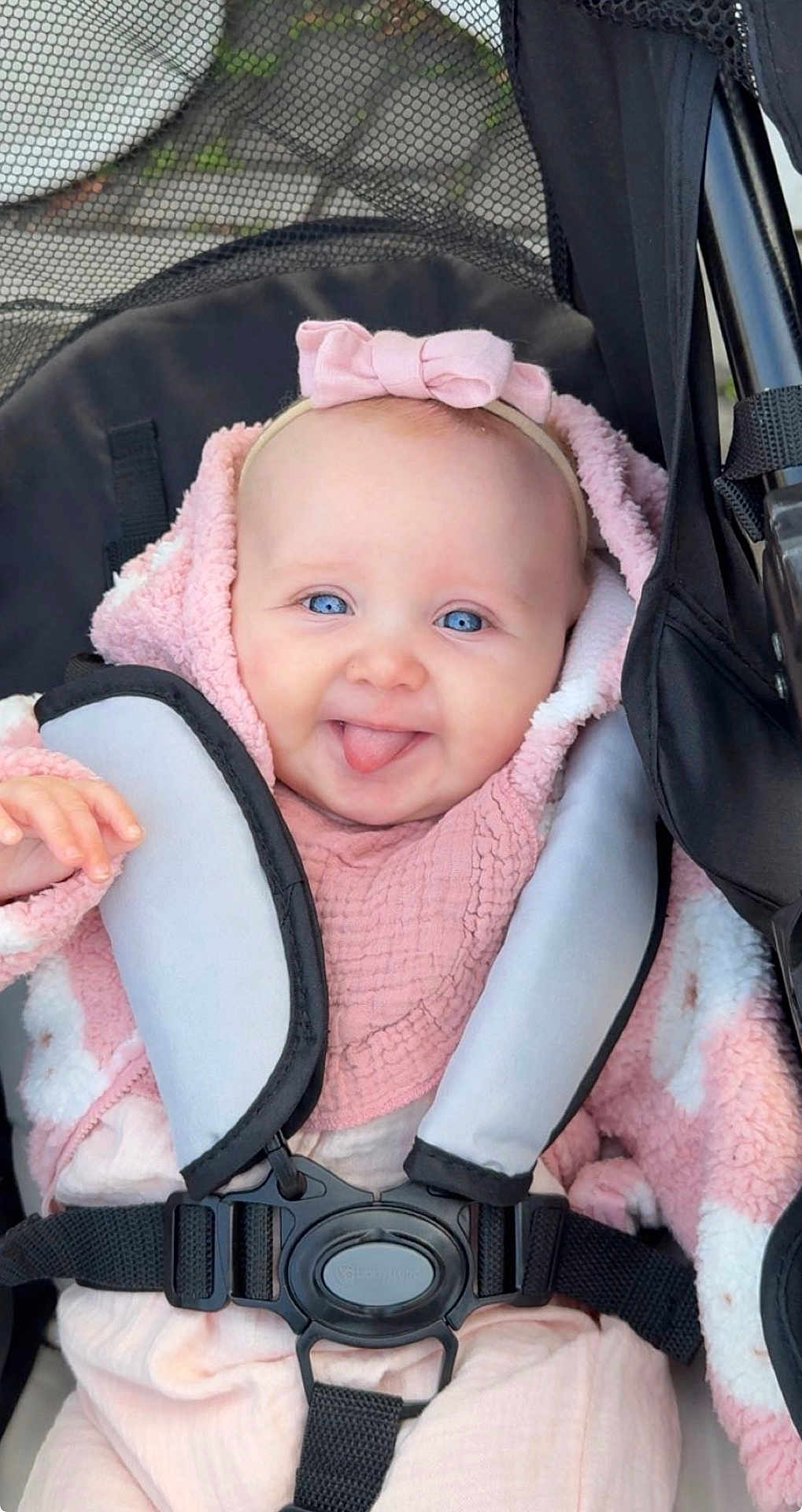 Aryah is registered to the contest to win money with this photo: baby, infant, stroller, car_seat, harness, strap, bow_headband, pink_clothing, blanket, blue_eyes, smile, tongue_out, cute, portrait, sitting, hoodie, plush, outdoor, canopy, closeup