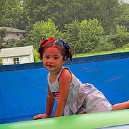 Harper is registered to the contest to win money with this photo: child, girl, curly_hair, bows, inflatable, playground, outdoor, trees, greenery, smiling, summer_clothing, tank_top, play, fun, candid, daylight, park, nature, happy, childhood