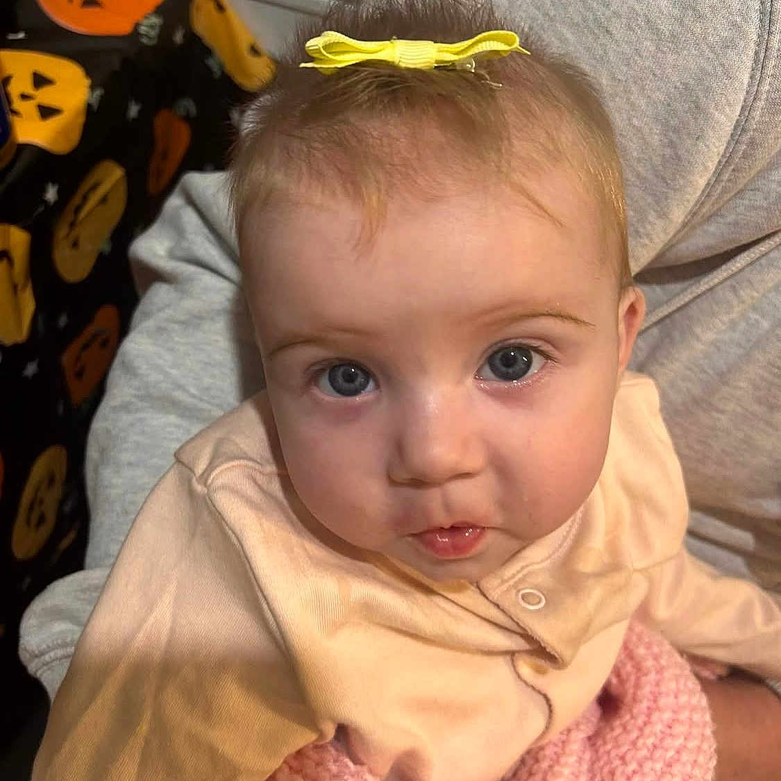 Dolcie is registered to the contest to win money with this photo: baby, blue_eyes, child, close_up, clothing, cute, fabric, gray_clothing, halloween, holding, indoor, infant, knitted_blanket, person, pink_blanket, portrait, pumpkin_pattern, sweatshirt, yellow_bow, young_child