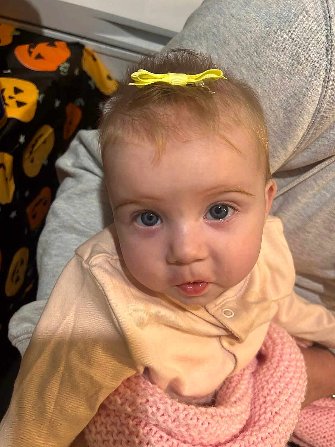 Dolcie is registered to the contest to win money with this photo: baby, blue_eyes, child, close_up, clothing, cute, fabric, gray_clothing, halloween, holding, indoor, infant, knitted_blanket, person, pink_blanket, portrait, pumpkin_pattern, sweatshirt, yellow_bow, young_child