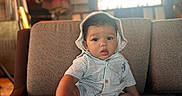 Asaiah joined the competition — help win amazing prizes! baby, child, infant, sitting, couch, indoor, hat, striped_clothing, white_socks, face, curious_expression, furniture, television, blurred_background, cozy, warm_lighting, person, portrait, young_child, home