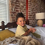 Asaiah is registered to the contest to win money with this photo: toddler, child, smiling, clapping, bed, blanket, pillow, yellow_shirt, curly_hair, brick_wall, window_blinds, lamp, wooden_headboard, indoors, happy, person, cozy, home, furniture, water_bottle
