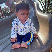 Liam is registered to the contest to win money with this photo: toddler, child, slide, playground, outdoor, sunlight, barefoot, tree, hand, clothing, pants, long_sleeve, expression, park, daylight, nature, play, young_child, casual, sitting