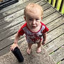 toddler, child, barefoot, carpet, porch, wooden_floor, red_shirt, minnie_mouse, tumbler, standing, looking_up, expression, hand, flag, outdoor, railing, clothing, person, daylight, grass