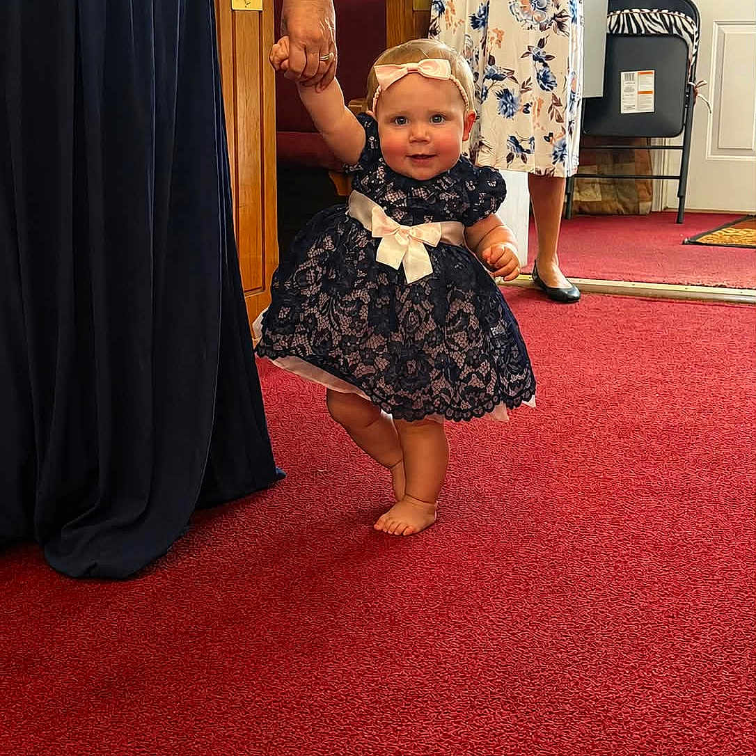 Charlie joined the competition — help win amazing prizes! baby, child, dress, lace, headband, bow, hand_holding, carpet, indoors, person, woman, footwear, barefoot, red_carpet, floral_dress, support, smiling, cute, celebration, event