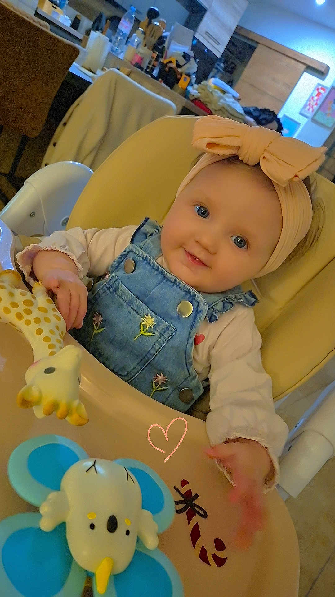 Livia participe au concours pour gagner de l'argent avec cette photo : baby, infant, highchair, headband, bow, denim_overalls, toy, giraffe_toy, elephant_toy, blue_eyes, smiling, rosy_cheeks, hand, feeding_table, indoor, kitchen, cozy, portrait, cute, doodle_heart