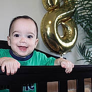 Eliott joined the competition — help win amazing prizes! baby, crib, balloon, number_six, green_shirt, smiling, happy, indoor, plant, black_crib, child, face, hands, home, celebration, birthday, infant, person, room, furniture