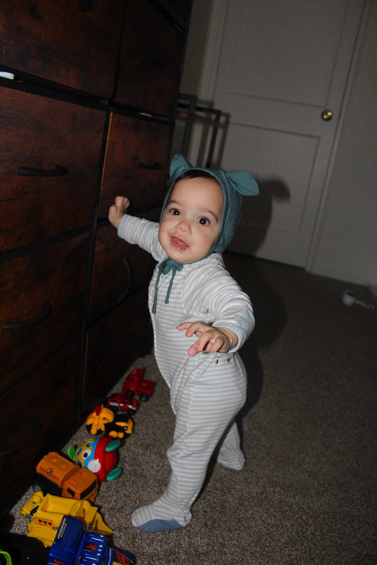 Eliott is registered to the contest to win money with this photo: toddler, child, toy, toys, car, cars, striped_pajamas, hat, blue_hat, ears, smiling, standing, carpet, dresser, wooden_dresser, indoor, room, happy, infant, play