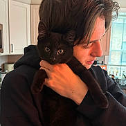 Kiko is registered to the contest to win money with this photo: person, cat, kitten, black_cat, portrait, indoor, kitchen, cupboard, window, holding, hug, young_person, face, fur, eyes, hoodie, home, cute, pet, close_up