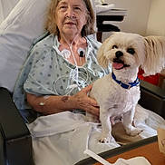 Chester joined the competition — help win amazing prizes! elderly_woman, hospital_bed, dog, small_dog, white_dog, hospital_gown, oxygen_tube, medical_equipment, hospital_room, bed, chair, table, plastic_fork, napkin, food_condiments, smiling_dog, pet, companion, indoor, patient