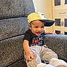 toddler, child, smiling, cap, baseball_cap, shirt, heart_graphic, pants, white_shoes, armchair, gray_furniture, indoor, happy, portrait, casual_clothing, young_child, cozy, seated, home, cute