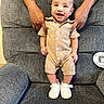 baby, smiling, romper, white_sneakers, tattooed_hands, gray_armchair, supporting_hands, happy, child, infant, seated, indoors, textured_clothing, cute, person, footwear, portrait, baby_fashion, comfortable, joyful