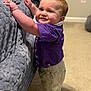 toddler, child, standing, purple_shirt, beige_shorts, carpet, bedspread, indoor, smile, face, hands, furniture, home, cute, baby, light, expression, person, young_child, portrait