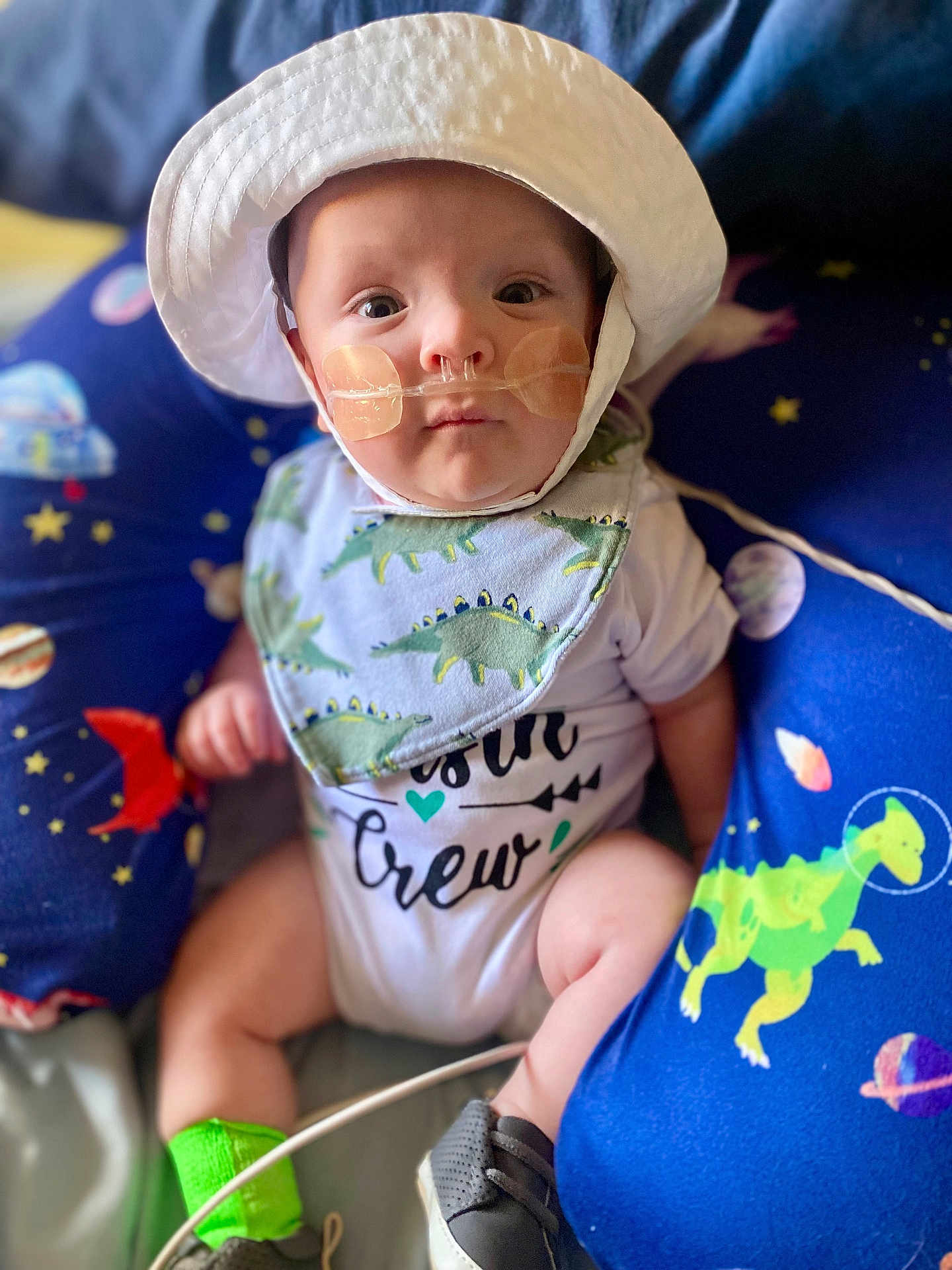 Calix joined the competition — help win amazing prizes! baby, infant, hat, bib, nasal_cannula, green_socks, dinosaur_pattern, space_theme, cushion, blue_background, white_clothing, close_up, indoors, seated, footwear, cute, child, portrait, medical_device, expression