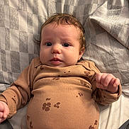 Elaina is registered to the contest to win money with this photo: baby, infant, child, face, person, clothing, brown_outfit, pattern, flower_pattern, blanket, checkered, bed, lying_down, cute, expression, portrait, indoors, soft_light, hair, fist