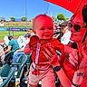 Oliver is registered to the contest to win money with this photo: baby, woman, umbrella, stadium, baseball, crowd, sunny, sunglasses, smile, red, shorts, shirt, hand, nails, bracelet, tattoo, watch, seating, outdoor, daytime