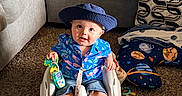 Oliver is registered to the contest to win money with this photo: baby, child, hat, blue_shirt, flamingo_pattern, baby_seat, toy, carpet, couch, pillow, patterned_pillow, living_room, floor, plush_toy, smiling, sitting, indoor, clothing, cute, person