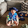 baby, child, hat, blue_shirt, flamingo_pattern, baby_seat, toy, carpet, couch, pillow, patterned_pillow, living_room, floor, plush_toy, smiling, sitting, indoor, clothing, cute, person