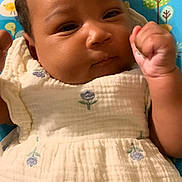 Kaliyah joined the competition — help win amazing prizes! baby, infant, child, close_up, face, hand, fist, dress, flower_pattern, white_clothing, curly_hair, indoor, baby_seat, colorful, blue, green, patterned, soft_texture, portrait, cute