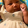baby, infant, child, close_up, face, hand, fist, dress, flower_pattern, white_clothing, curly_hair, indoor, baby_seat, colorful, blue, green, patterned, soft_texture, portrait, cute