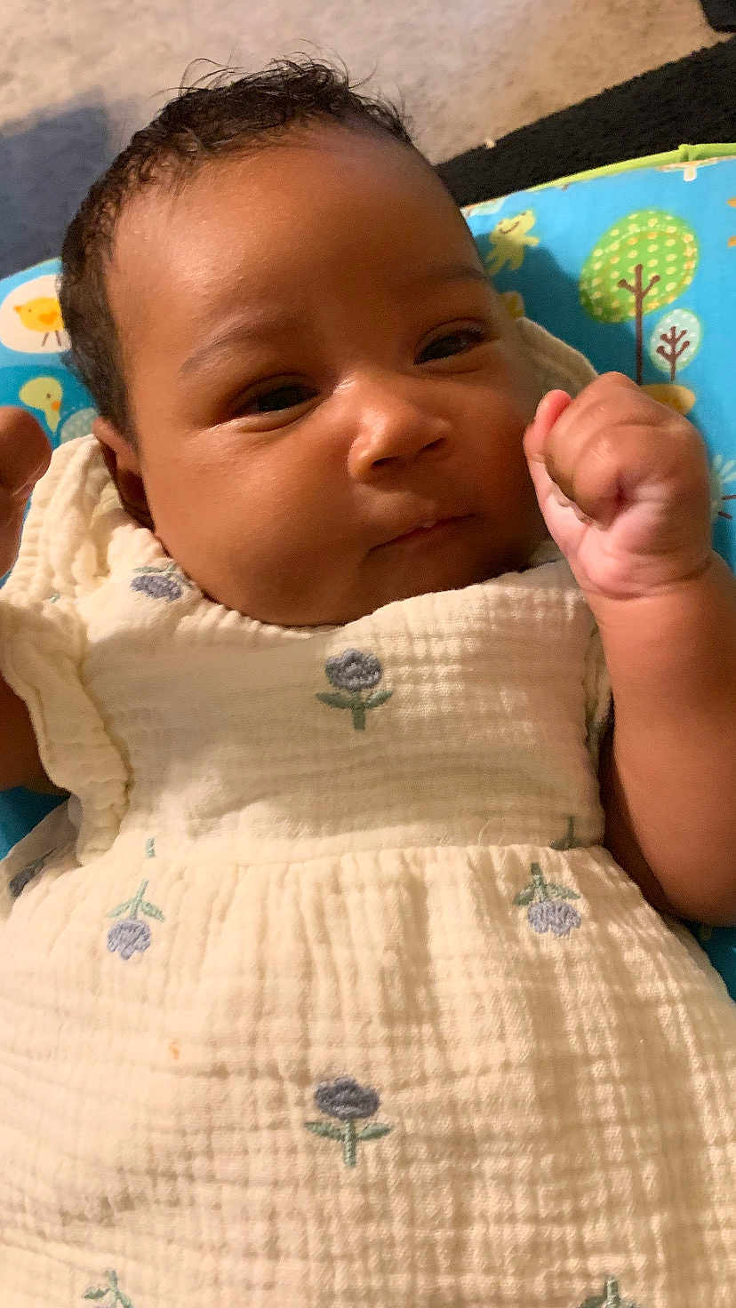 Kaliyah joined the competition — help win amazing prizes! baby, infant, child, close_up, face, hand, fist, dress, flower_pattern, white_clothing, curly_hair, indoor, baby_seat, colorful, blue, green, patterned, soft_texture, portrait, cute
