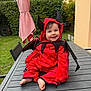 Mira participe au concours pour gagner de l'argent avec cette photo : baby, child, costume, devil, red, outdoor, table, smiling, face, happy, greenery, umbrella, garden, feet, clothing, person, cute, sitting, playful, hood