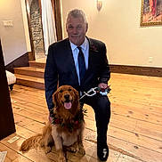 Lea is registered to the contest to win money with this photo: man, dog, golden_retriever, indoor, wooden_floor, suit, tie, flower_wreath, pet, smiling_dog, portrait, stairs, wall_art, lamp, happy, kneeling, brown_dog, decor, leash, wooden_stairs