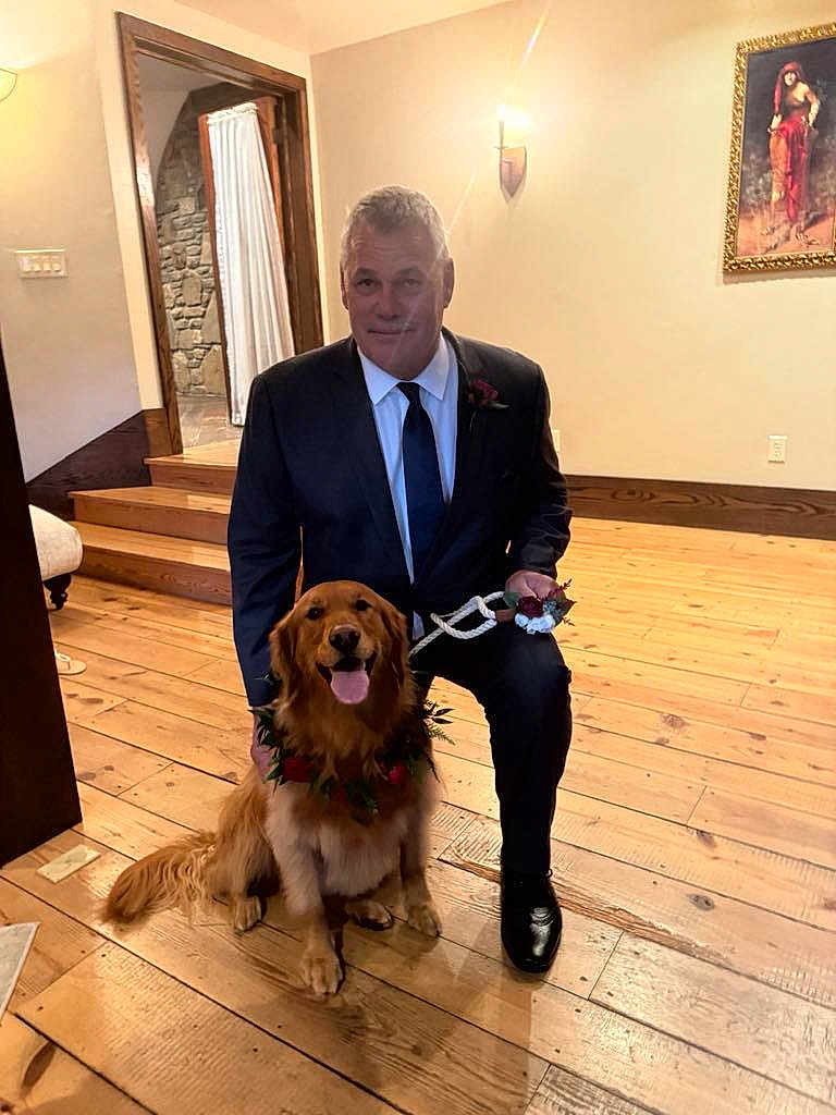 Lea is registered to the contest to win money with this photo: man, dog, golden_retriever, indoor, wooden_floor, suit, tie, flower_wreath, pet, smiling_dog, portrait, stairs, wall_art, lamp, happy, kneeling, brown_dog, decor, leash, wooden_stairs