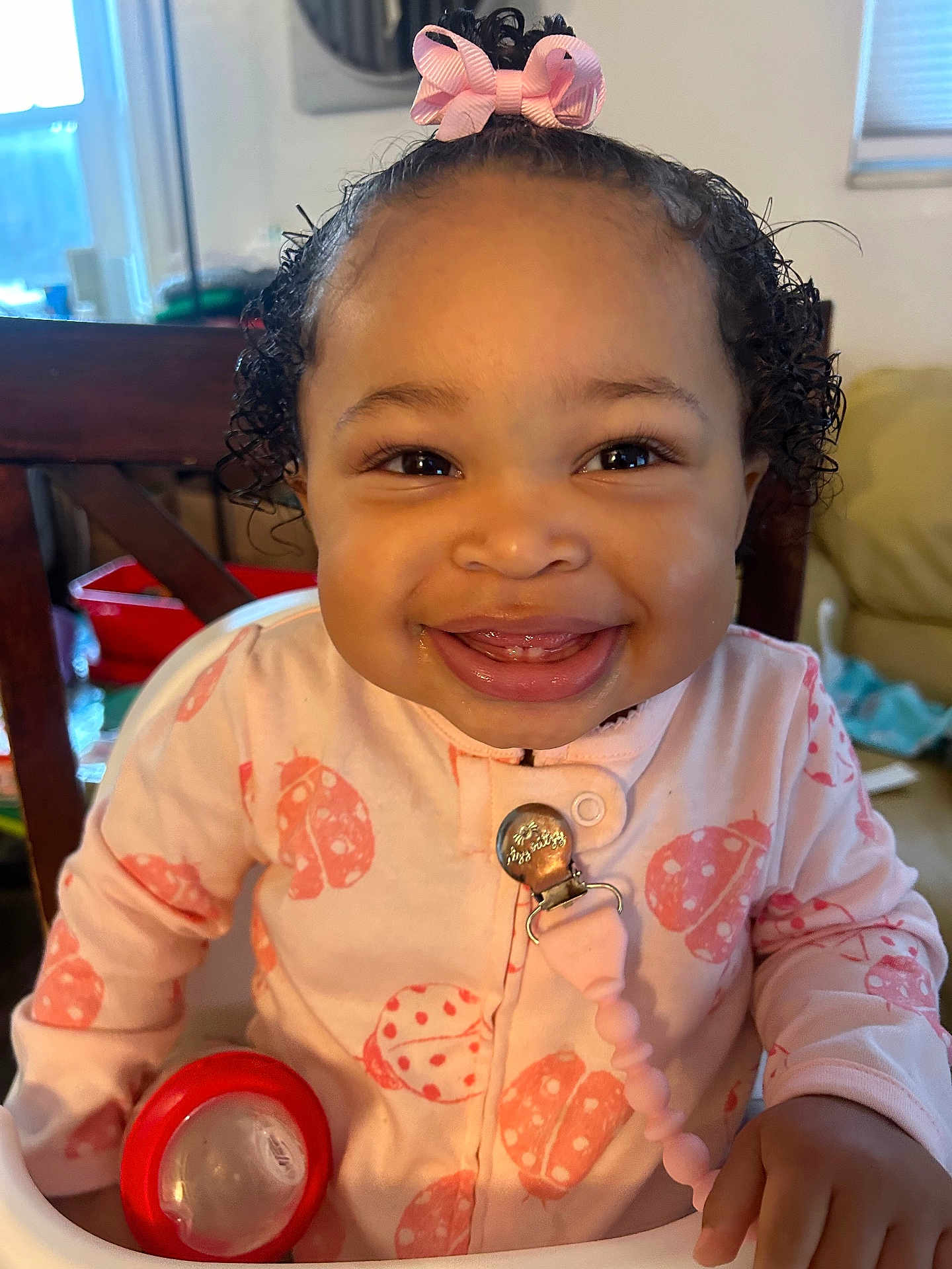 Zennayah joined the competition — help win amazing prizes! baby, smiling, child, high_chair, pink_bow, curly_hair, onesie, ladybug_pattern, red_cup, indoors, furniture, person, happy, cute, toddler, home, seat, clothing, face, portrait