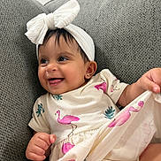 Innie is registered to the contest to win money with this photo: baby, child, smiling, headband, bow, flamingo_print, dress, couch, grey, happy, cute, infant, clothing, playful, person, portrait, indoors, fabric, hand, sitting