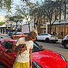 person, dog, car, sports_car, street, sidewalk, tree, building, sky, cloud, urban, vehicle, casual_clothing, yellow_shorts, white_tshirt, red_car, daytime, outdoor, pet, holding