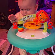 John is registered to the contest to win money with this photo: baby, child, indoor, walker, toy, lion_toy, carpet, colorful, playtime, footwear, person, floor, lighting, furniture, activity, cute, infant, exploring, room, play