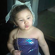 Zara is registered to the contest to win money with this photo: toddler, child, girl, curly_hair, bow, mermaid_outfit, purple_dress, shiny_clothing, necklace, puckered_lips, indoor, cute, portrait, baby, fashion, costume, playful, young_child, person, headwear